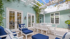 Une terrasse couverte avec des meubles blancs et des coussins bleus, une terrasse en bois et une maison d’un bleu clair.