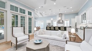 Un salon spacieux avec un grand canapé blanc, deux fauteuils blancs et une table basse. La pièce est dotée de grandes fenêtres, d’un ventilateur de plafond et d’un éclairage encastré.