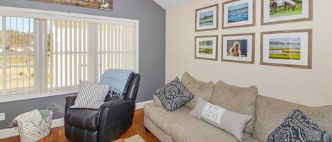 Een woonkamer met een grijze bank, een zwarte leren fauteuil en een houten bordje met de tekst 'THE SEA HORSE'. Er hangen vier ingelijste foto's aan de muur.