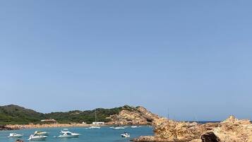 A coastal scene with rocky terrain, clear blue water, and several boats anchored near the shore.