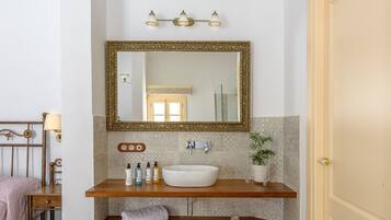 A bathroom with a large mirror, a white sink, and a wooden vanity. There are toiletries on the vanity and a towel hanging on the wall.