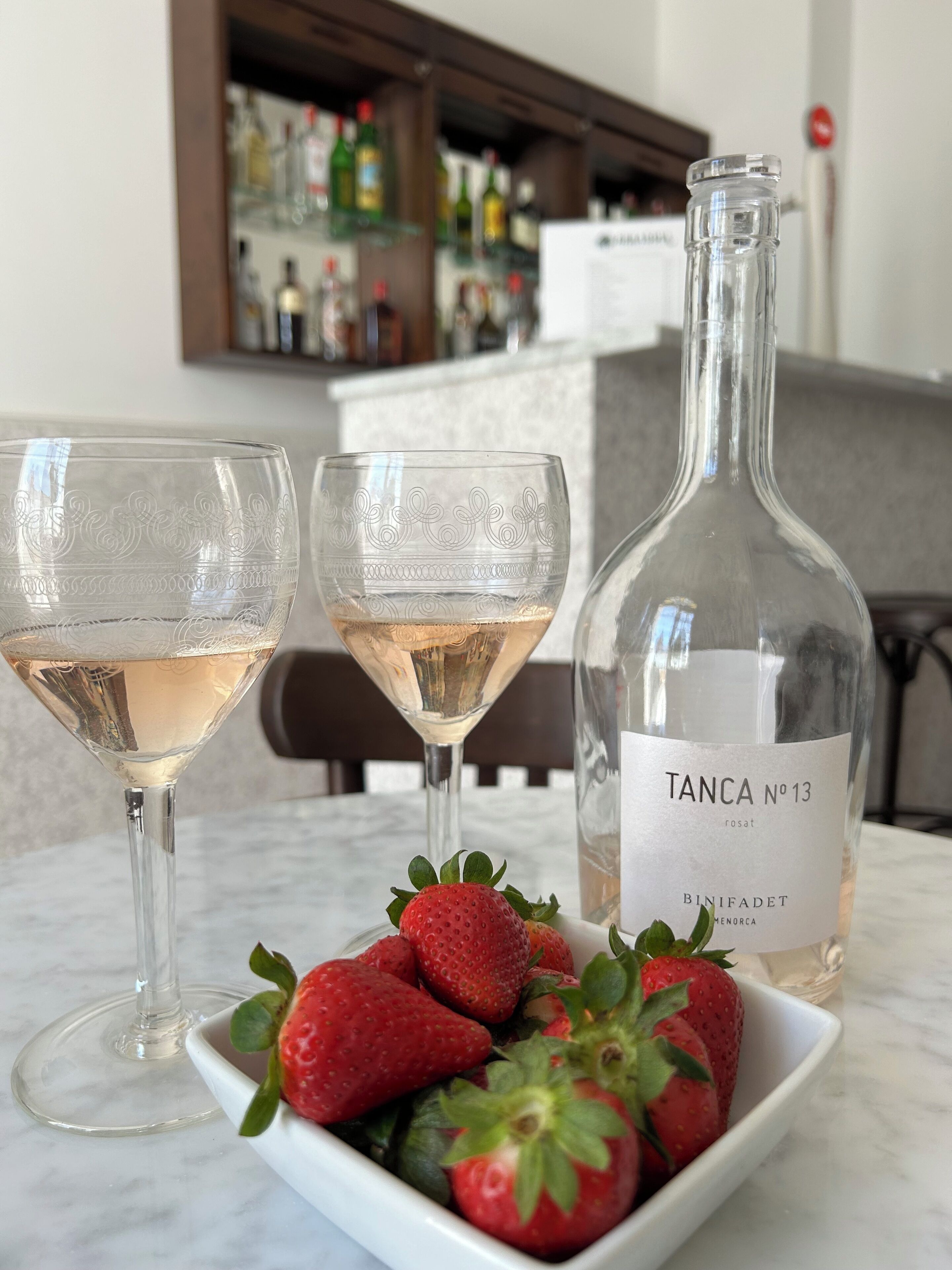 Dos copas de vino rosado, una botella de Tanca No. 13 y un bol de fresas sobre una mesa de mármol.