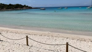 Una playa con aguas turquesas cristalinas, una orilla arenosa y varios barcos anclados en la distancia.