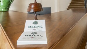 Mostrador de recepción en el Hotel Serranova Luxury, con superficie de madera, un jarrón verde con flores, una campana y un cartel.