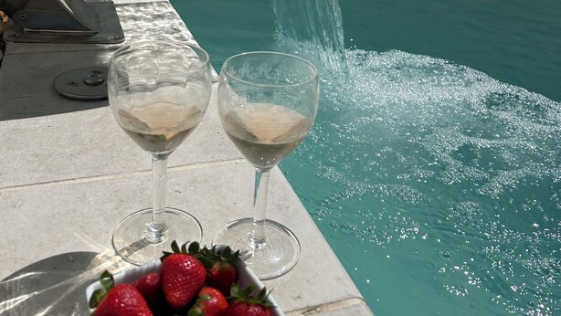 Escena junto a la piscina con fuente de agua, dos copas de vino blanco y un bol de fresas.