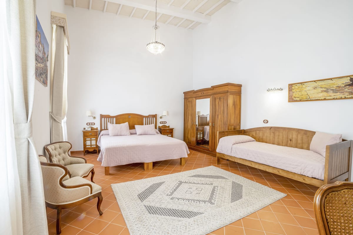 Ein Schlafzimmer mit einem Bett, zwei Stühlen, einem Sofa, einem Holzschrank, einem Spiegel und einer Pendelleuchte.