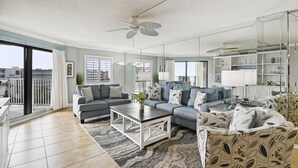 A spacious living room with a grey sofa set, a coffee table, and a patterned armchair. There is a large window with blinds, a ceiling fan, and a mirrored wall reflecting the room. - Inlet Reef 618 is Recently Upgraded 2 BR 2 Ba on the beach - amazing sunset views! by RedAwning (Destin)