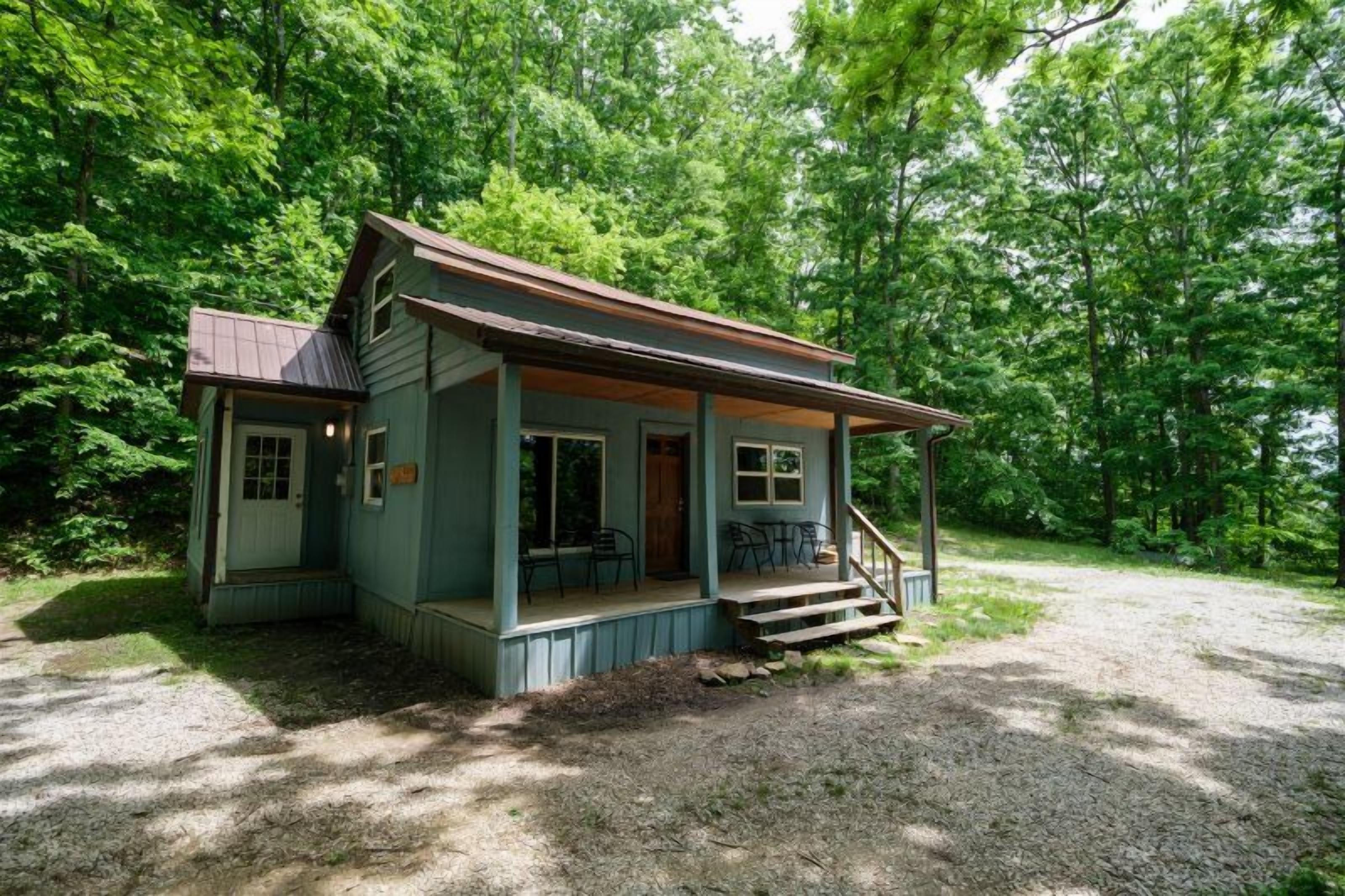 Une petite cabane à un seul étage, dotée d’un porche et d’une allée en gravier, entourée d’arbres denses.