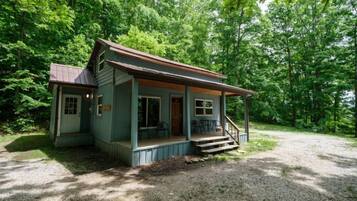 Une petite cabane à un seul étage, dotée d’un porche et d’une allée en gravier, entourée d’arbres denses.
