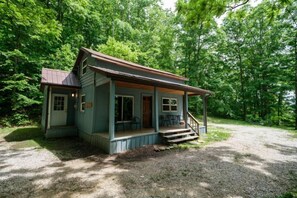 A small, single-story cabin with a porch and a gravel driveway, surrounded by dense trees.