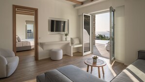 A modern living room with a sofa, ottoman, and a small table with a plant. A TV is mounted on the wall, and there's a sliding glass door leading to an outdoor area. - Arocaria (Mykonos)