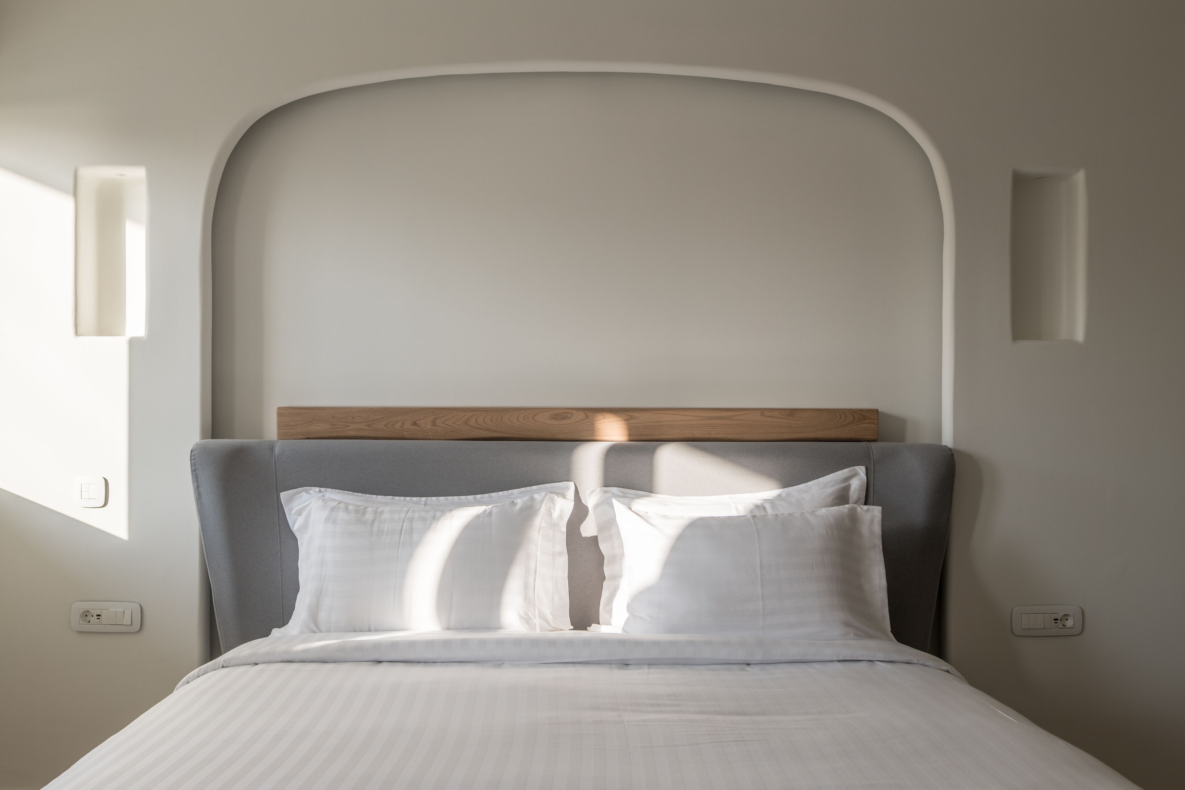 Un lit bien fait, avec du linge de lit blanc et des oreillers, une tête de lit en bois et un mur de couleur claire agrémenté d’un plafond en retrait.