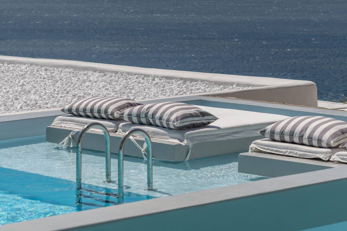 A rooftop infinity pool with lounge chairs and pillows, overlooking a body of water.