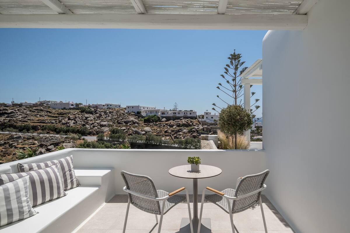 A balcony with a white sofa, two wicker chairs, and a small table with a plant. A rocky landscape and buildings are visible in the background.