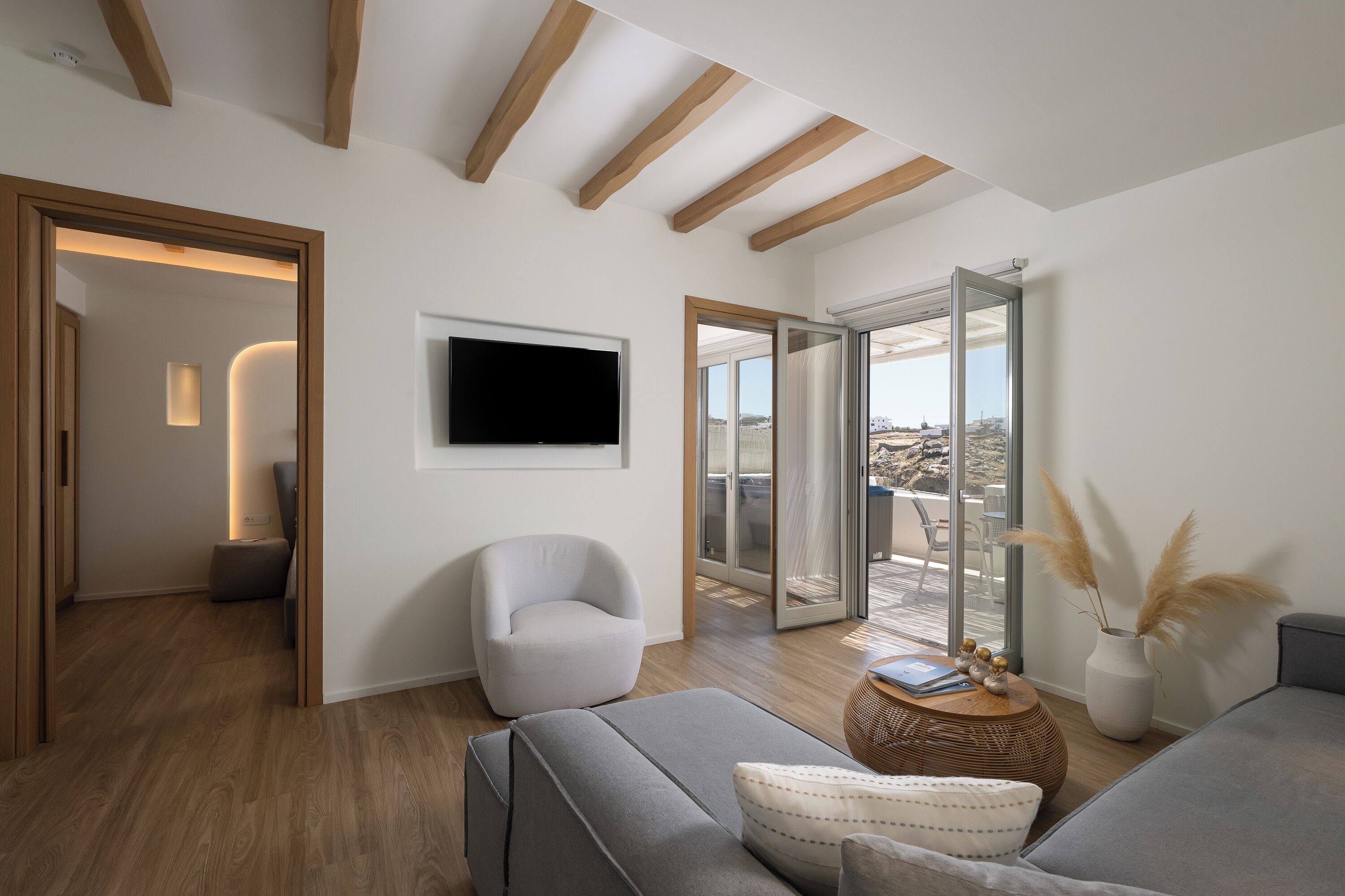 A modern living room with a sofa, armchair, flat-screen TV, and a view through glass doors to an outdoor area.