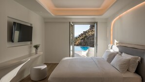 A modern bedroom with a large bed, a TV mounted on the wall, and a view of a pool and rocky landscape through an open door. - Arocaria (Mykonos)