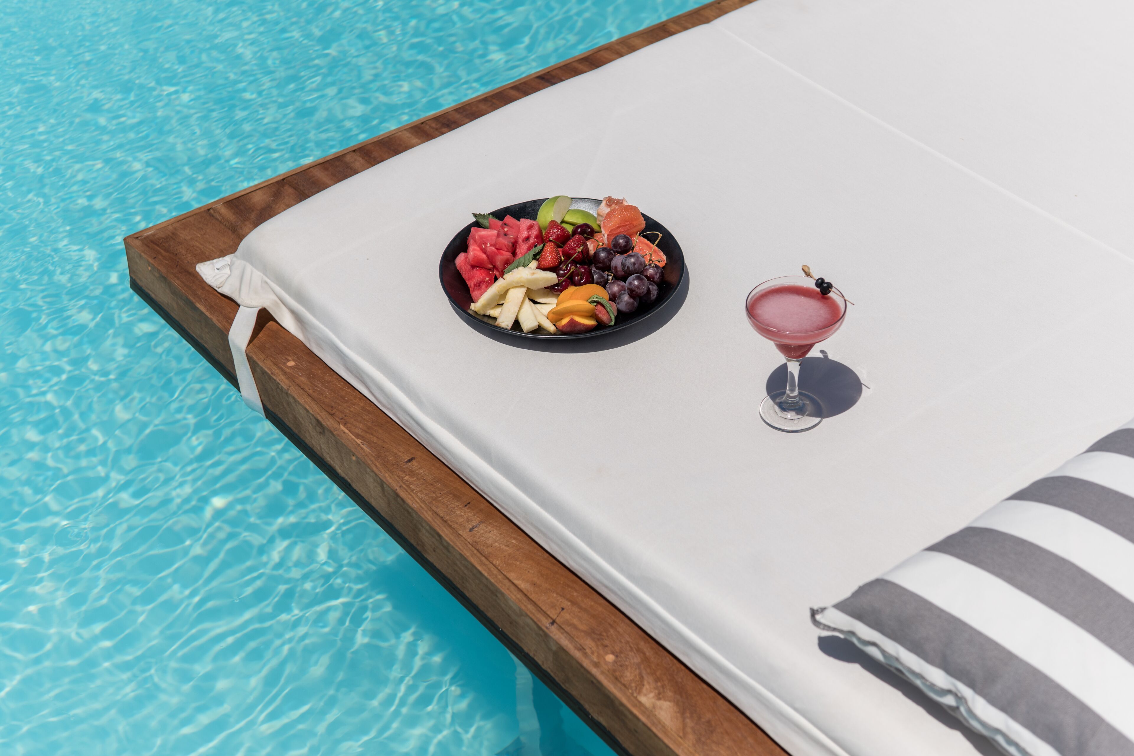 Escena junto a la piscina con cubierta de madera, una silla de descanso blanca, una bandeja de frutas y una copa de cóctel.