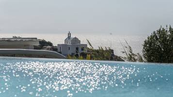 A pool with clear water reflecting sunlight, a white building with a dome and a bell tower, and a view of the sea with a distant ship.
