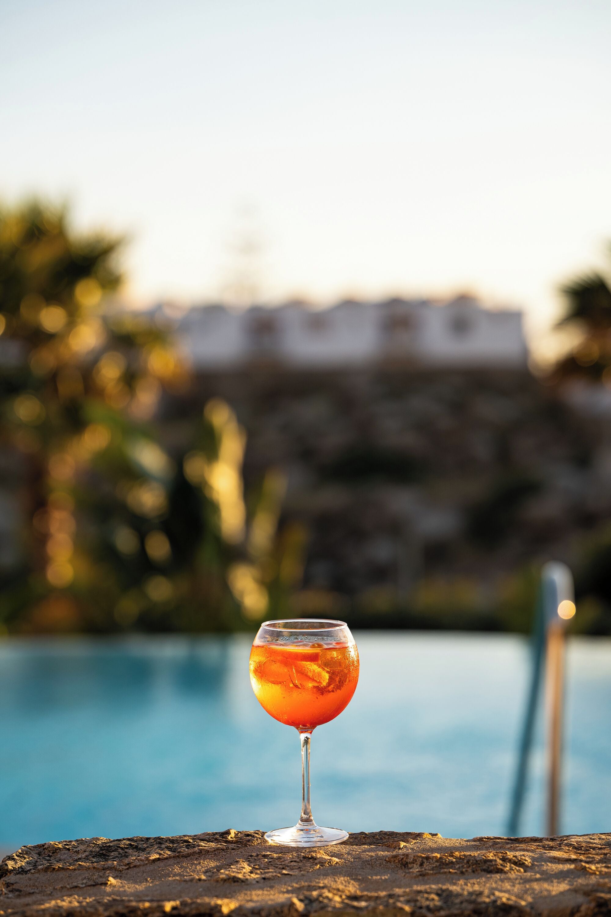 Un verre de boisson à l’orange sur une table près d’une piscine, avec un arrière-plan flou de bâtiments et de verdure.