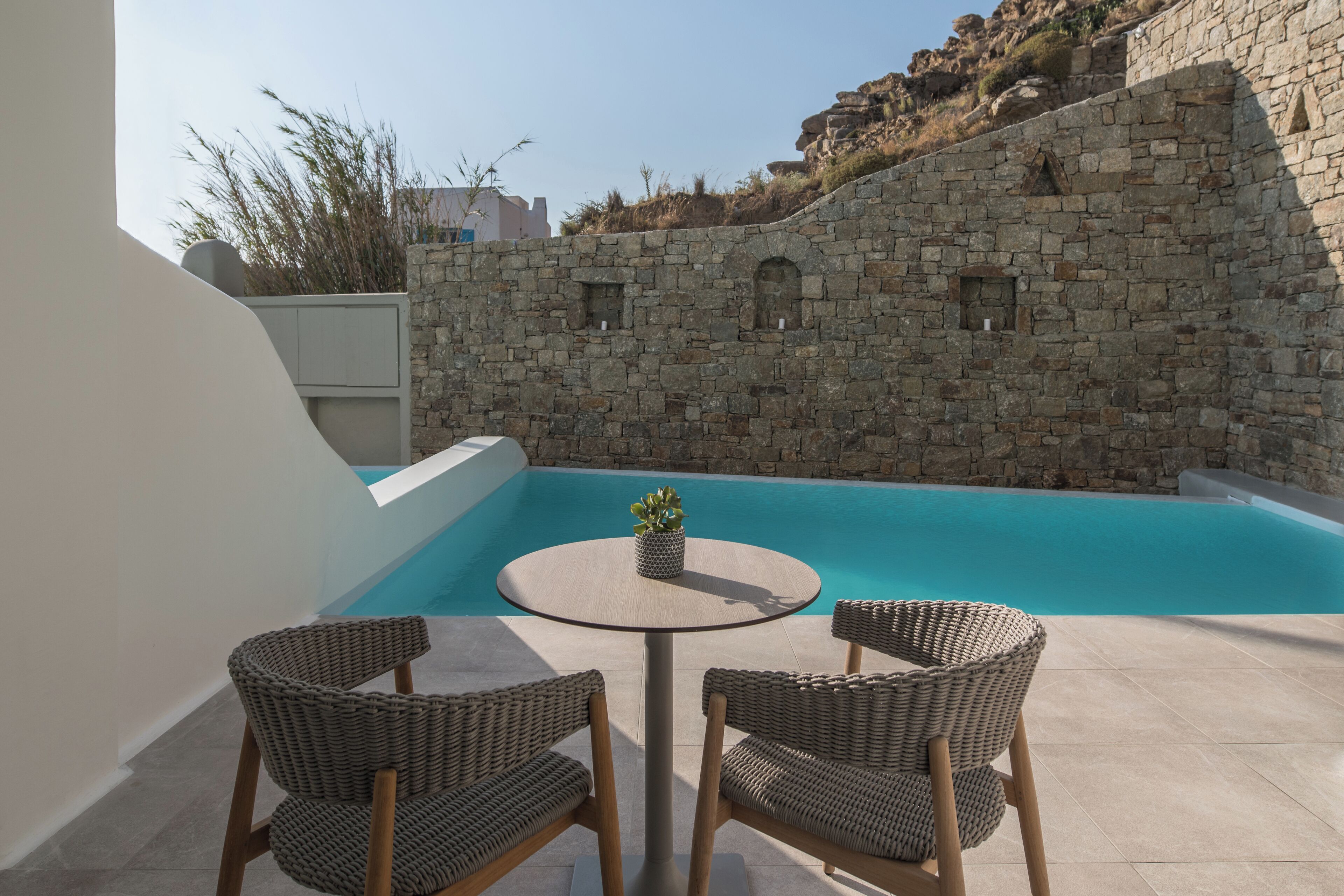 A poolside area with a round table and wicker chairs, a stone wall, and a clear blue pool.