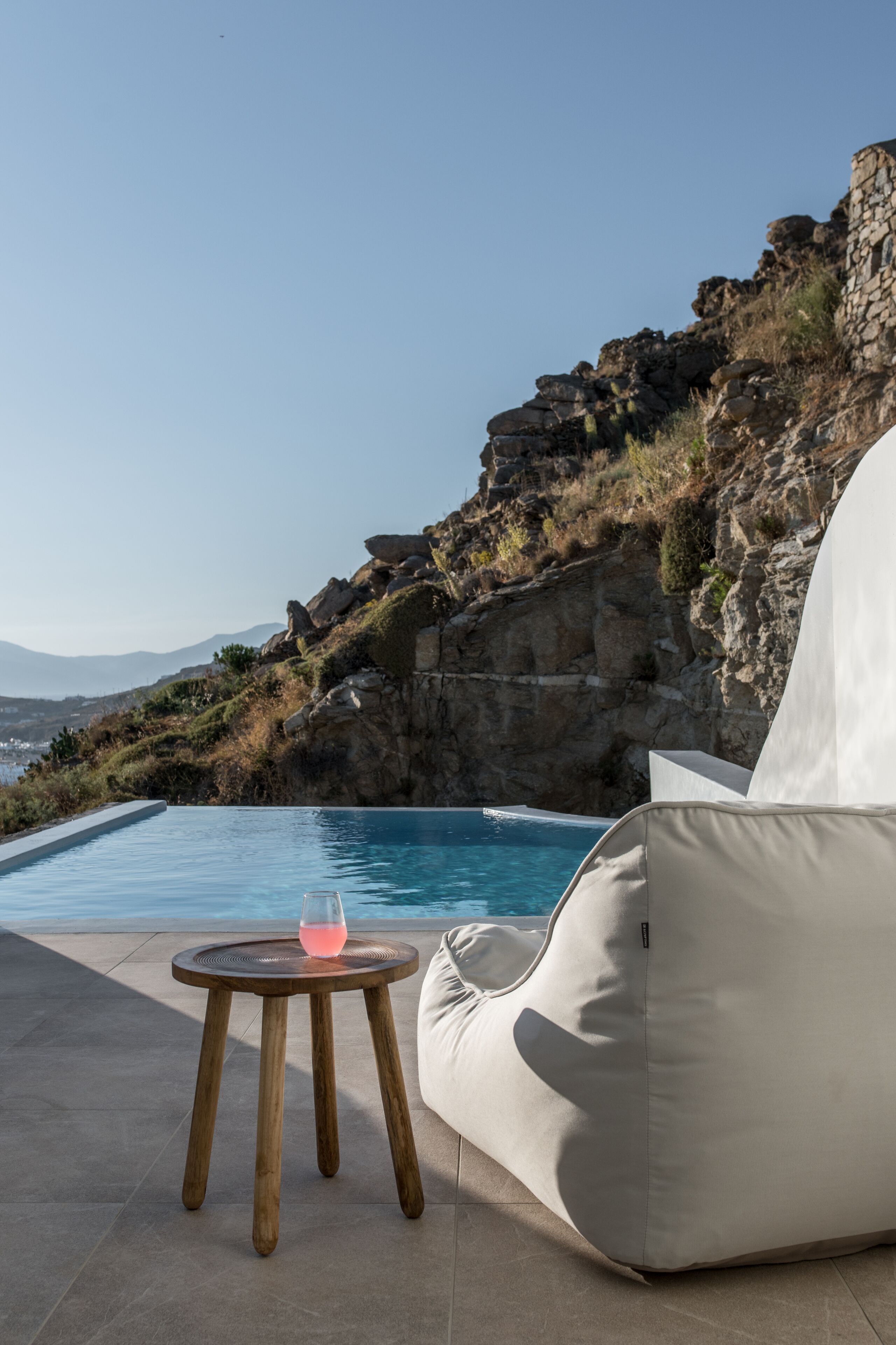 A poolside area with a white lounge chair, a small round table with a pink drink, and a rocky cliff in the background.