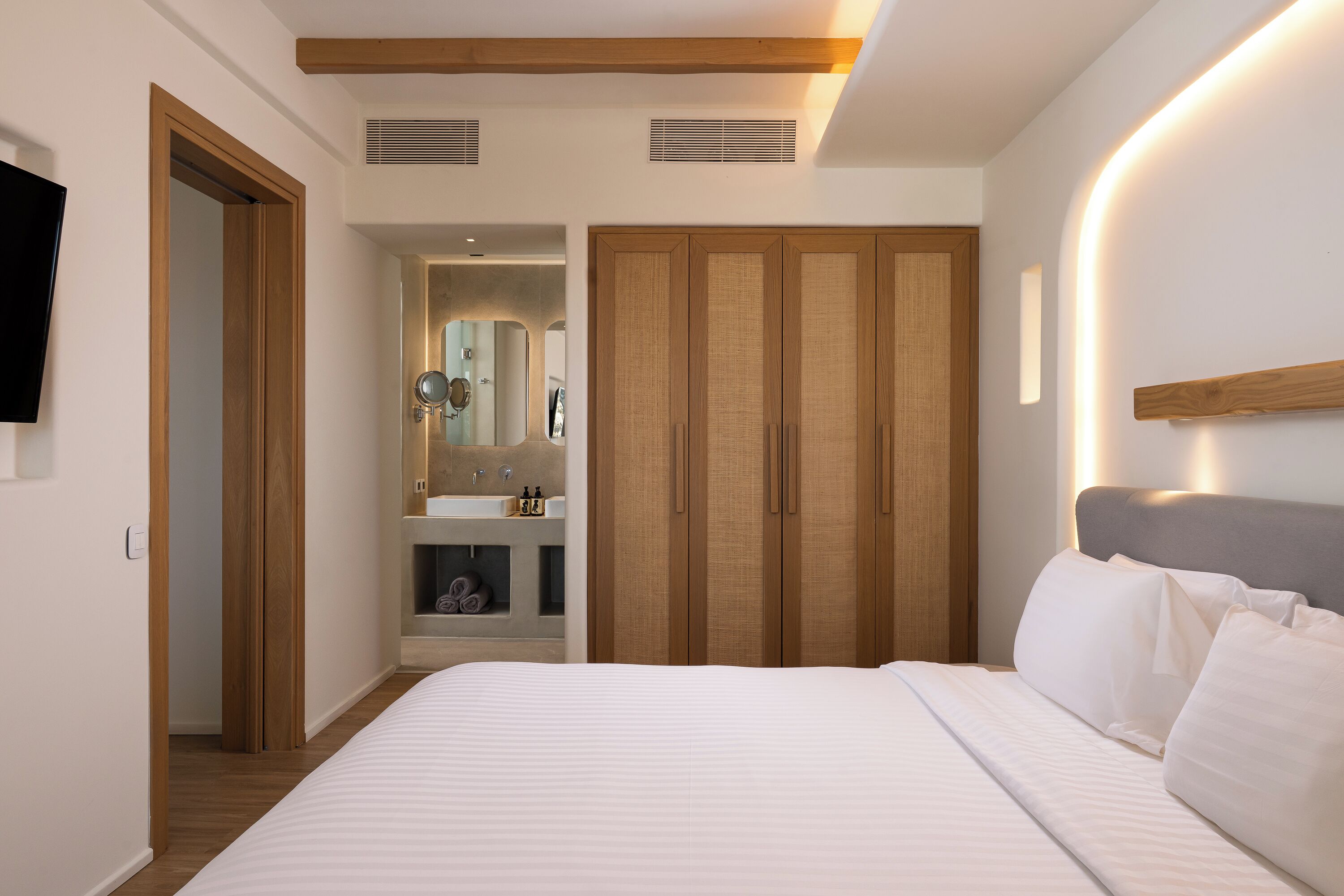 Een slaapkamer met een bed, een houten garderobe, een badkamer met spiegel en een televisie aan de muur.