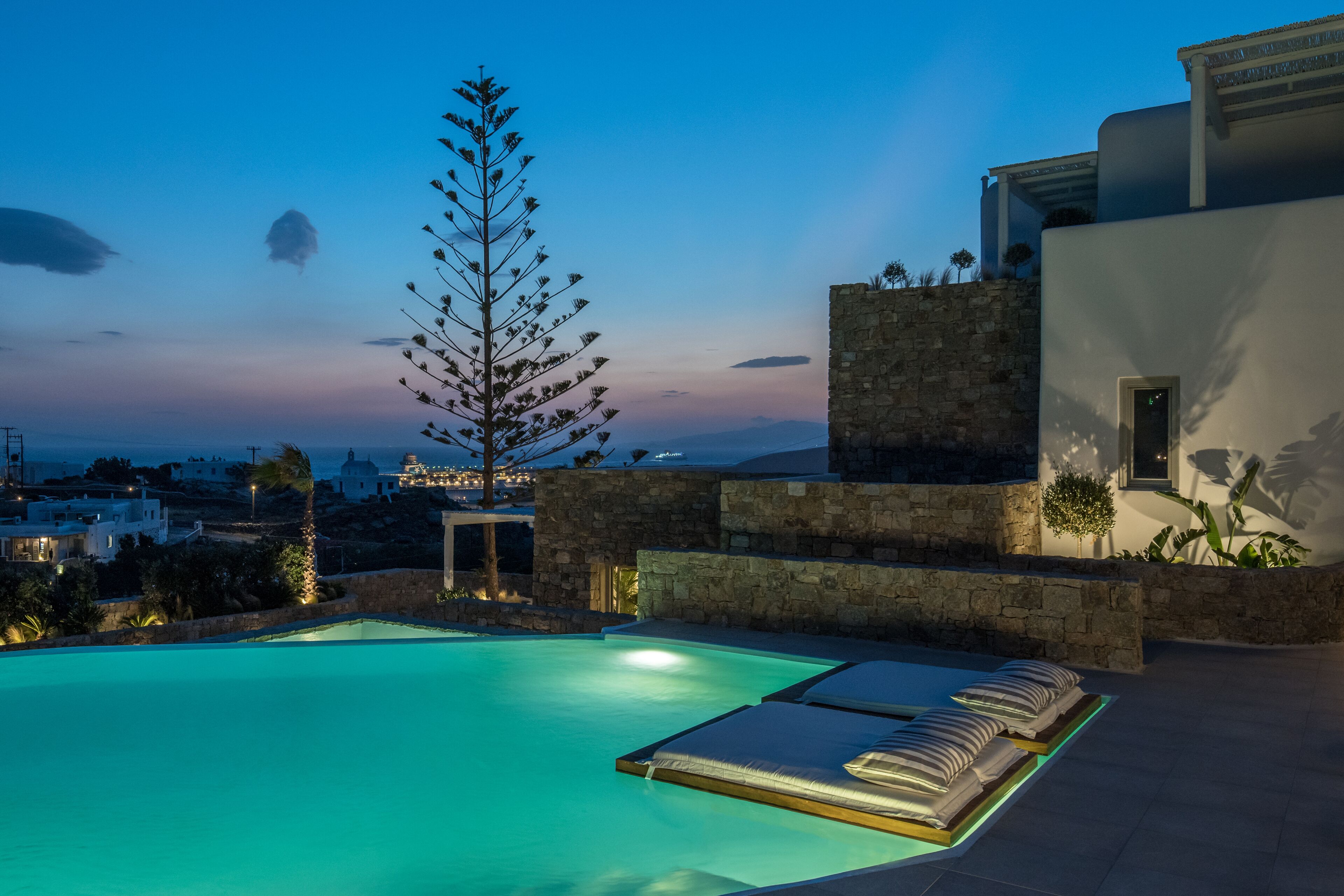 A pool area with lounge chairs and a stone wall, overlooking a cityscape at dusk.