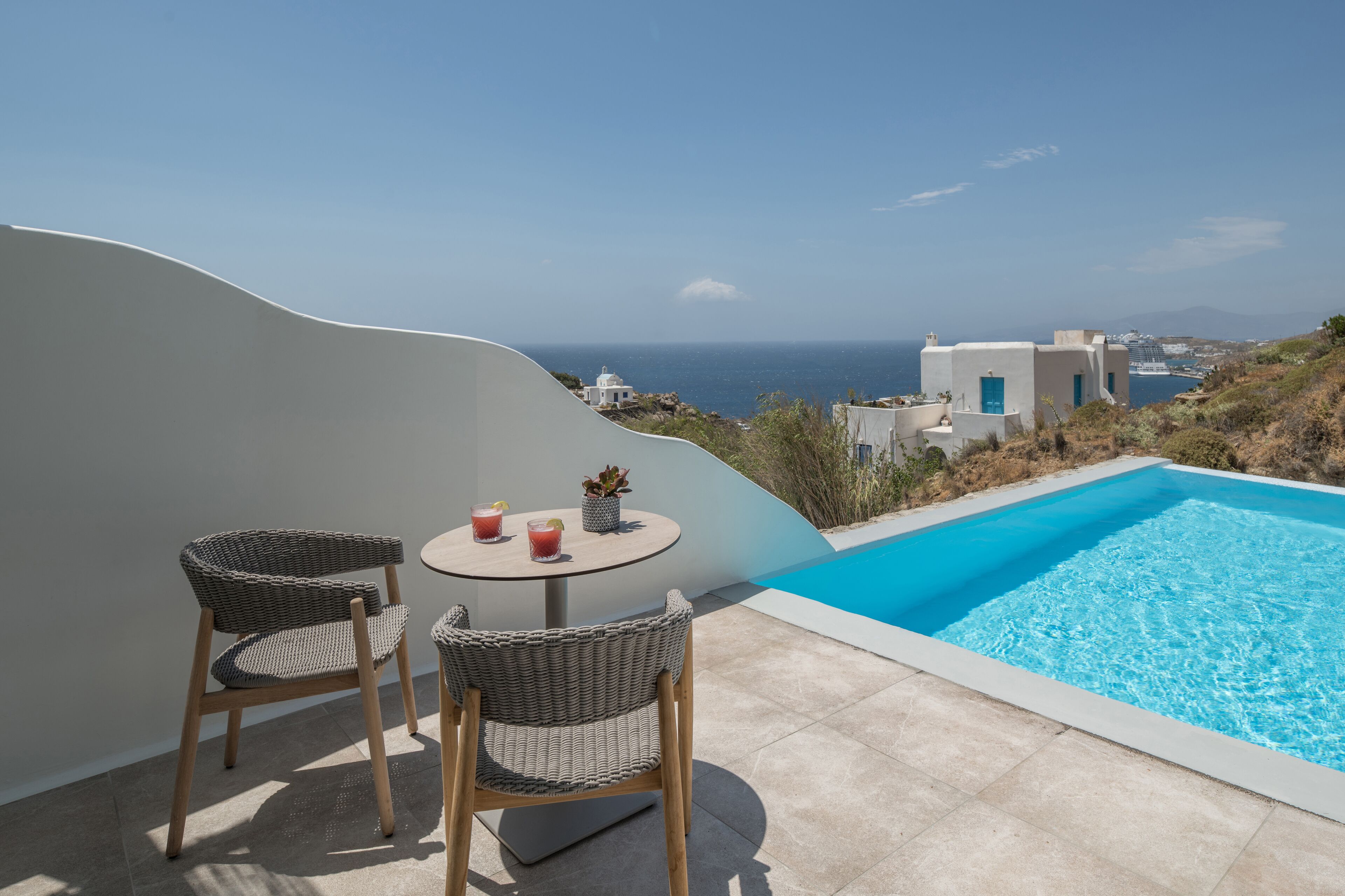 A poolside area with two wicker chairs and a table, overlooking a clear blue sea and white buildings.