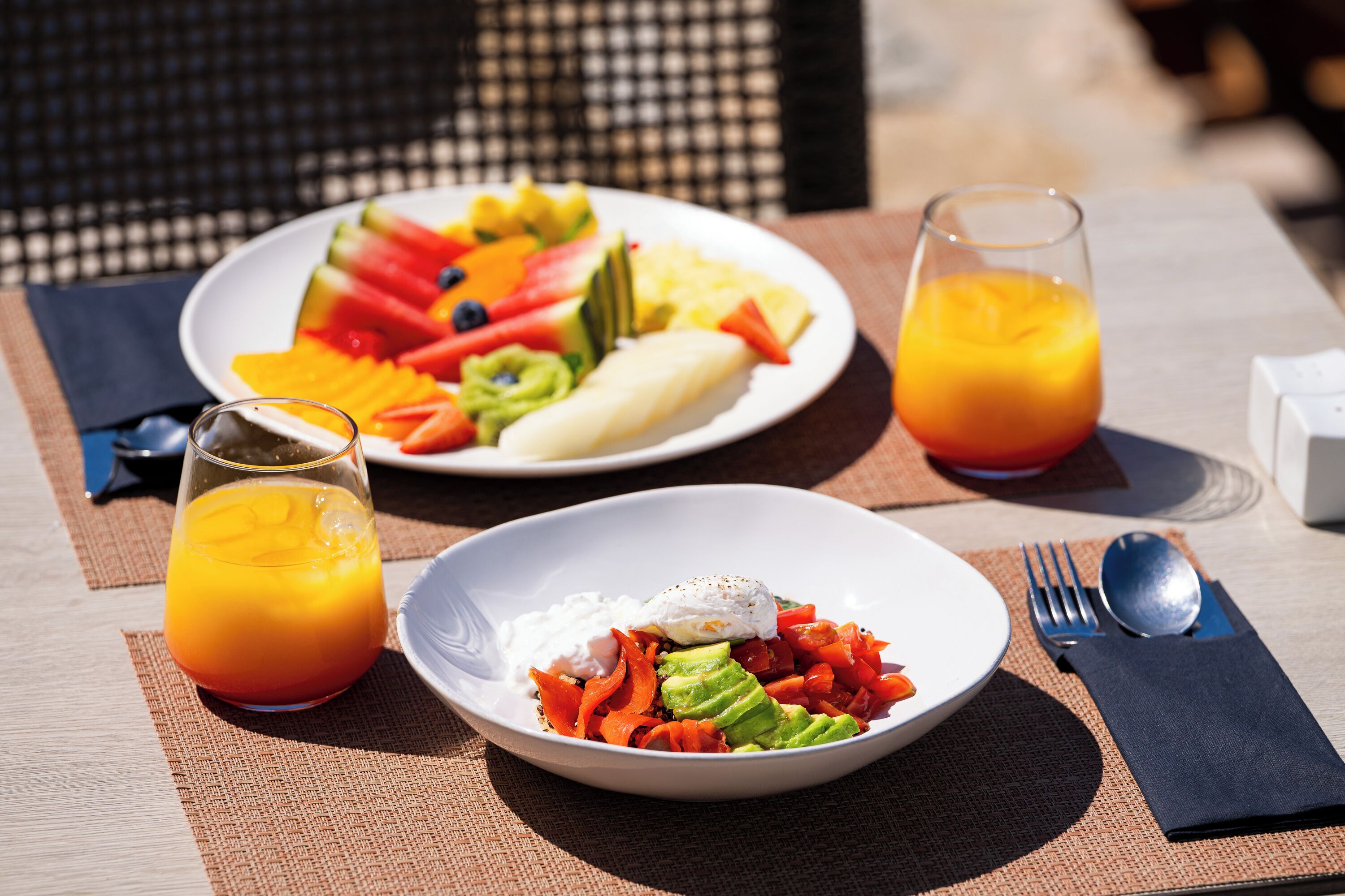 Una mesa con platos de fruta, aguacate y huevos, un vaso de zumo de naranja, y un tenedor y una cuchara.