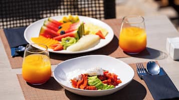 A breakfast setting with plates of fruit, avocado, and eggs, a glass of orange juice, and a fork and spoon on a table.