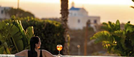 A person in a pool with a drink, surrounded by greenery and a building in the background.