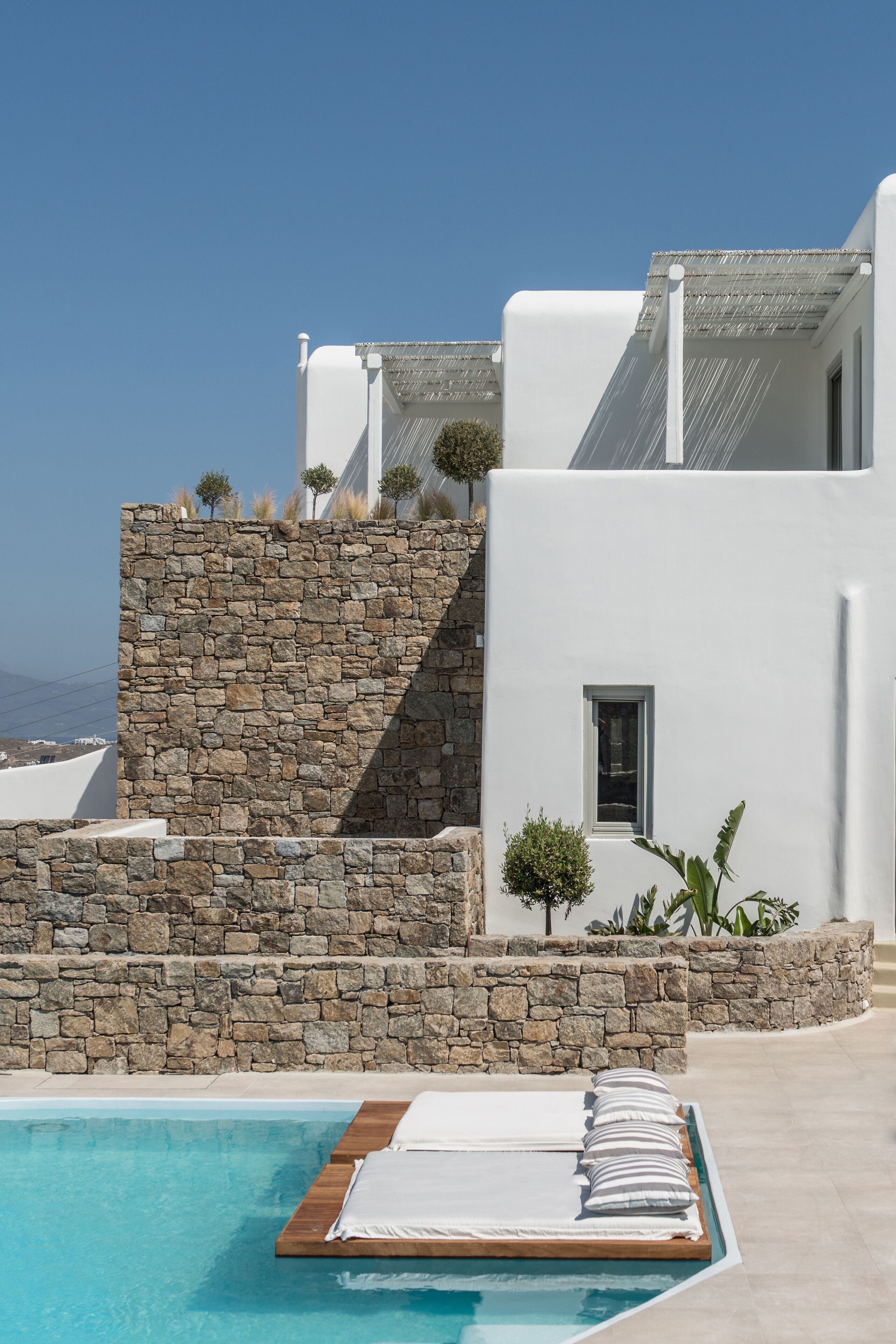 A modern building with a stone wall and a pool area with a wooden deck and white lounge chairs.