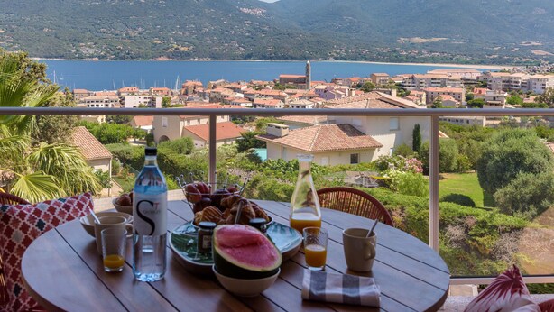 A table with breakfast items, a bottle, and glasses, overlooking a coastal town with mountains in the background. - Deluxe sea view apartment (Propriano)