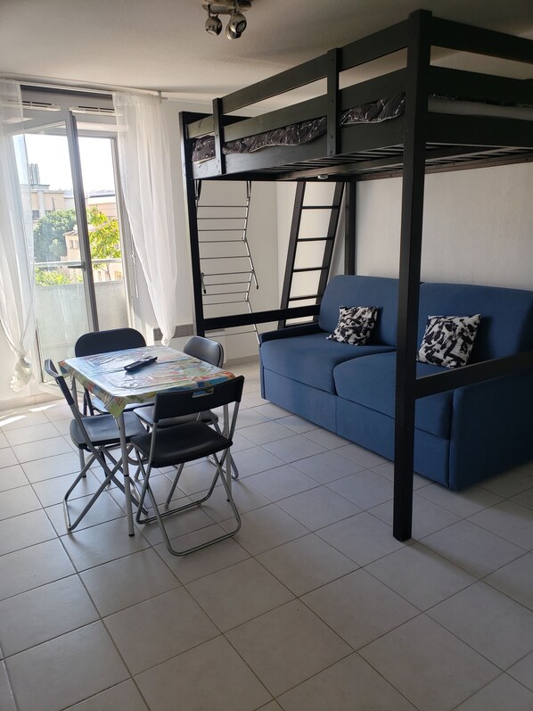 A room with a bunk bed, a blue sofa with patterned cushions, a dining table with chairs, and a view of the outside through a window.