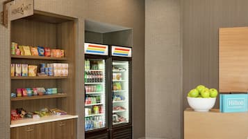 A hotel room with a minibar, snack shelves, a refrigerator, and a bowl of apples on a wooden table.