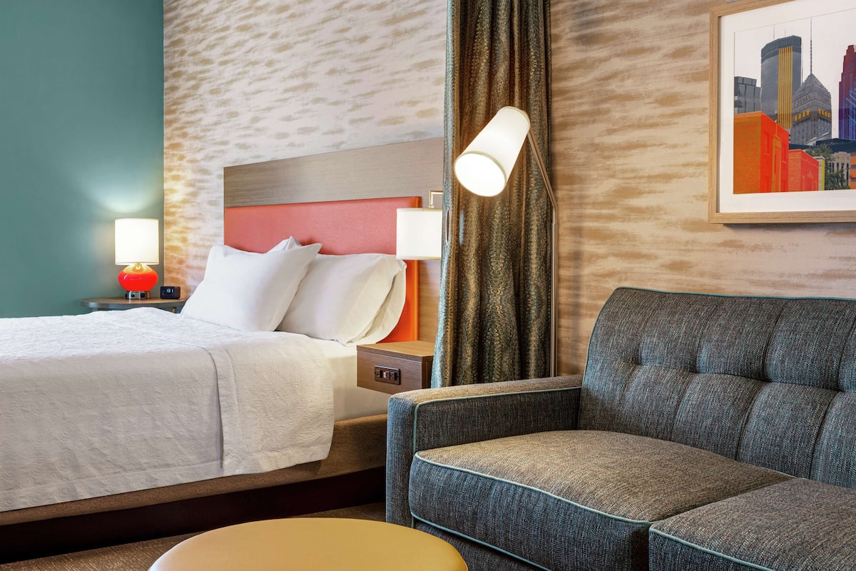 Een hotelkamer met een bed, een slaapbank, een nachtkastje met een lamp, en een lijst met een stadsgezicht aan de muur.