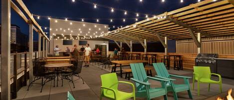 An outdoor patio area with wooden tables and chairs, string lights, and a fire pit.