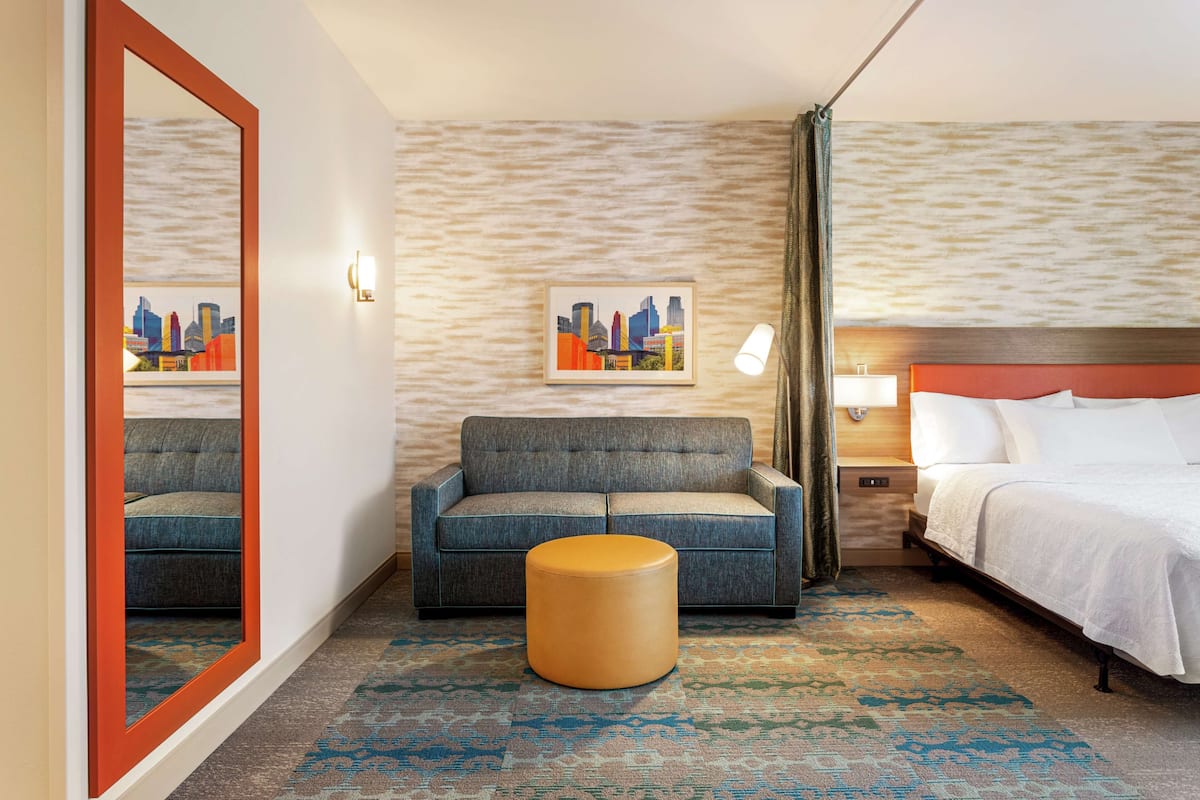 Een hotelkamer met een bed, een sofa, een rond poef, een spiegel en twee stadsgezicht schilderijen aan de muur.