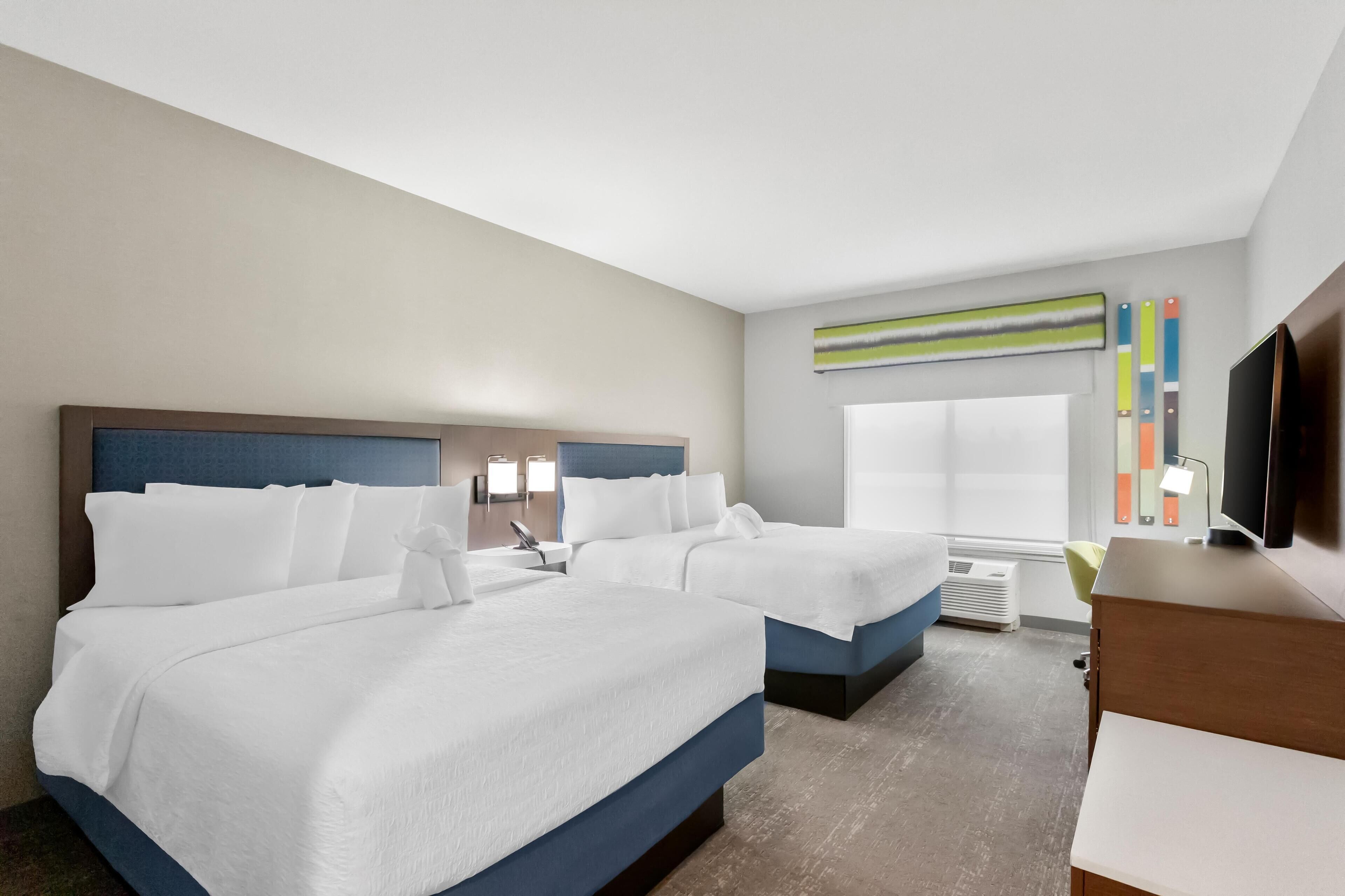 A hotel room with two beds, a TV, a desk, and a window with blinds.