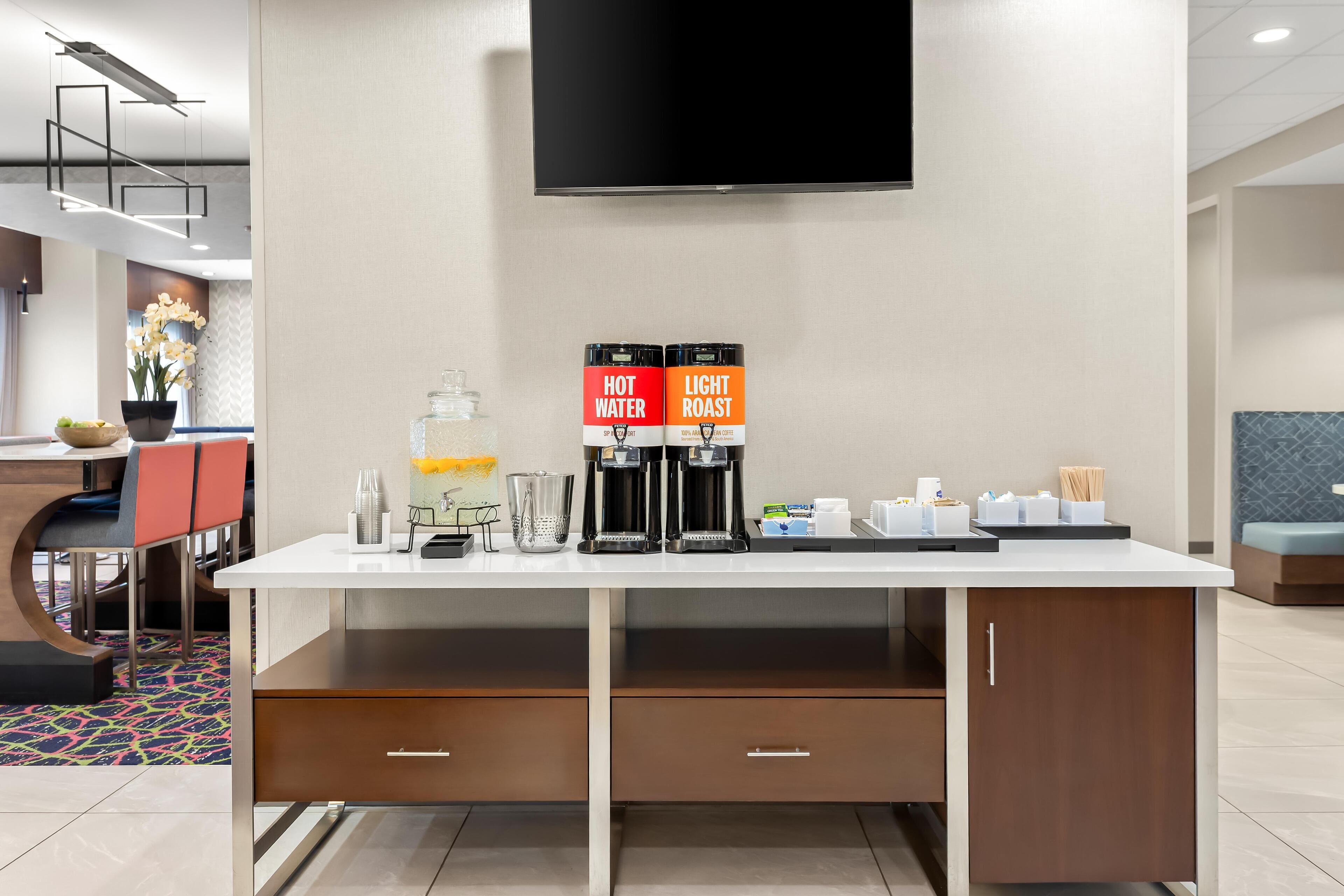 A hotel reception area with a coffee station, a television mounted on the wall, and a seating area with a patterned rug.