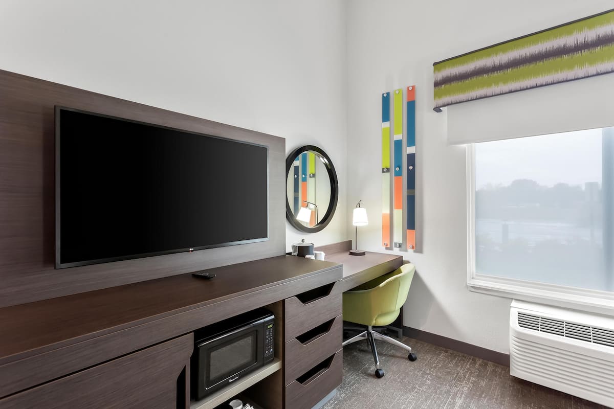 Quarto de hotel com uma grande televisão de tela plana, micro-ondas, escrivaninha com cadeira, espelho redondo e janela com vista para a cidade.