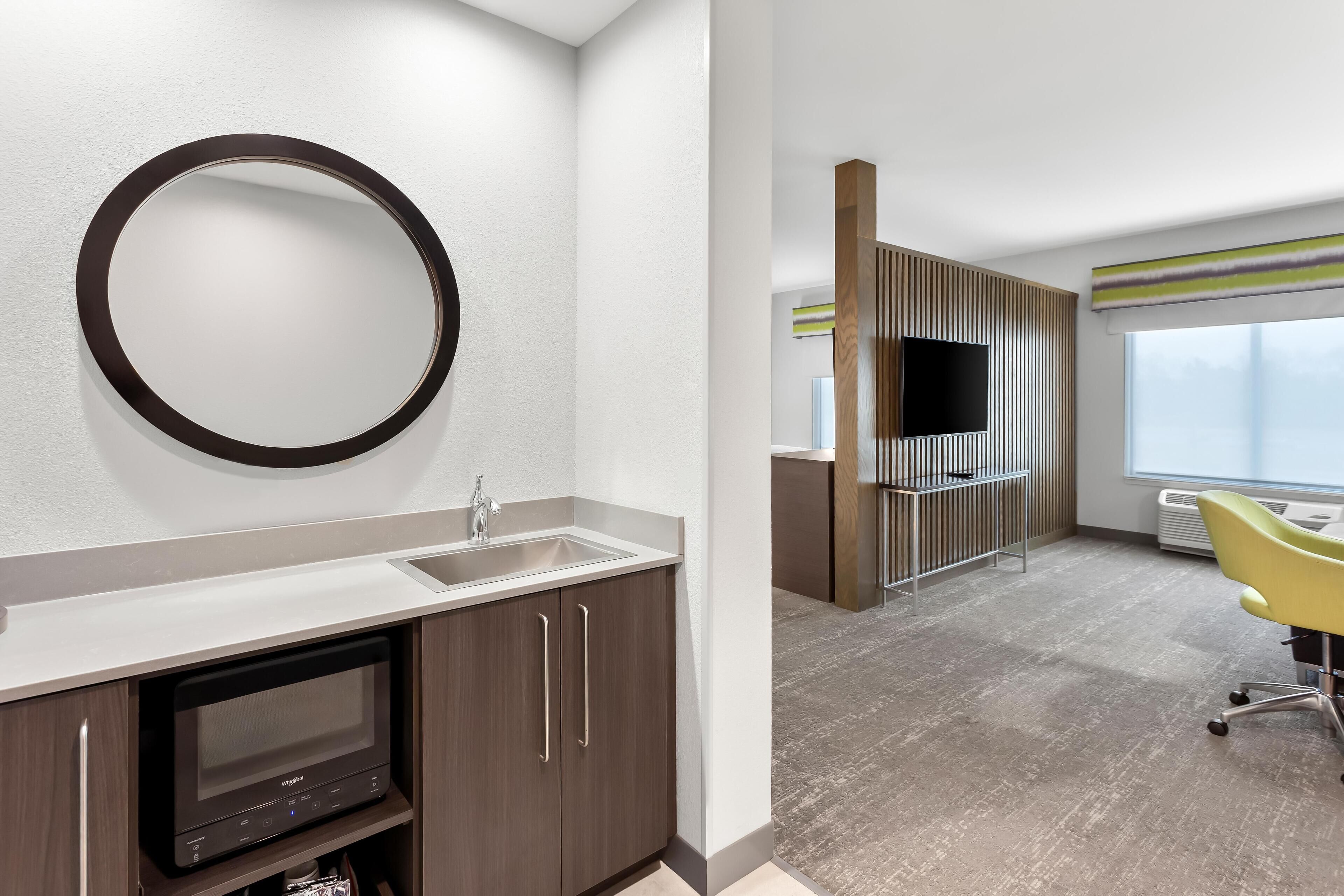 A modern hotel room with a large mirror, a built-in TV, a sink, and a yellow chair.