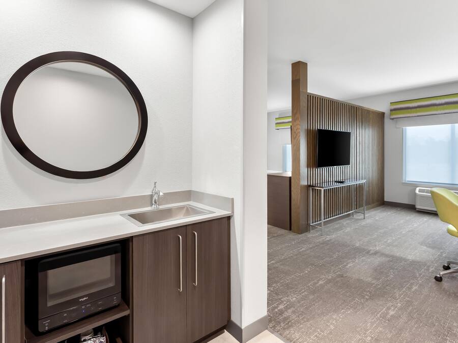 A modern hotel room with a large mirror, a built-in TV, a sink, and a yellow chair.