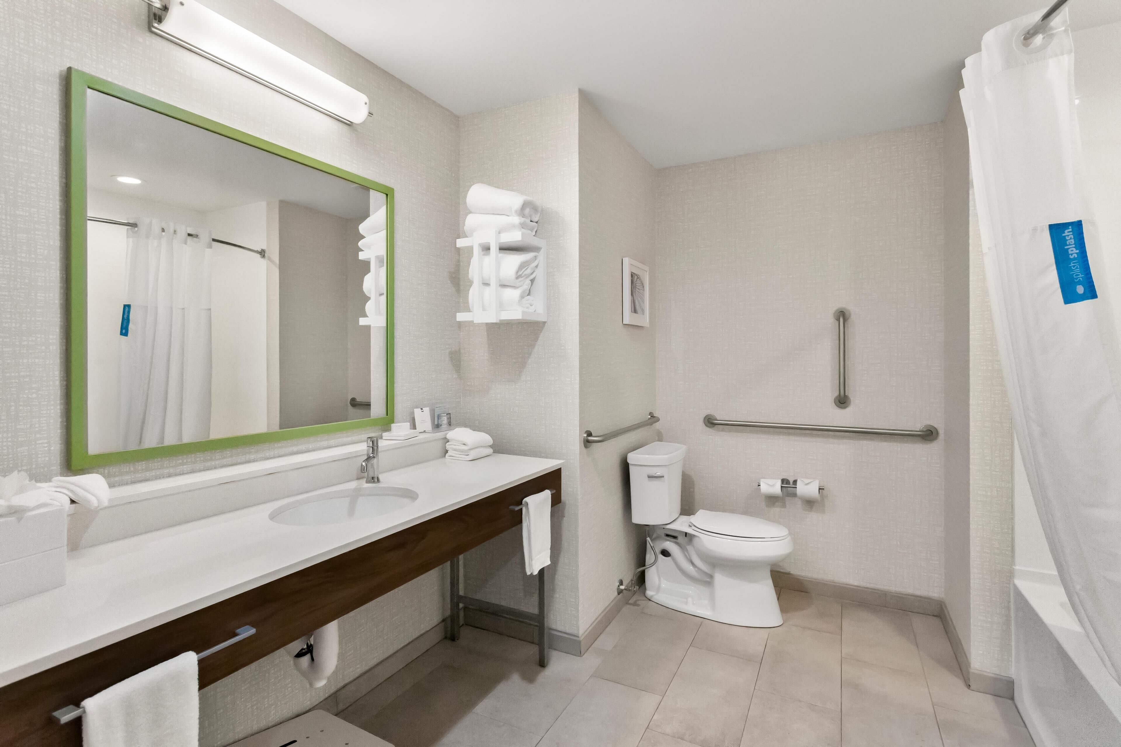 A modern bathroom with a dual sink vanity, a toilet, a shower with a grab bar, and a mirror with a green frame.