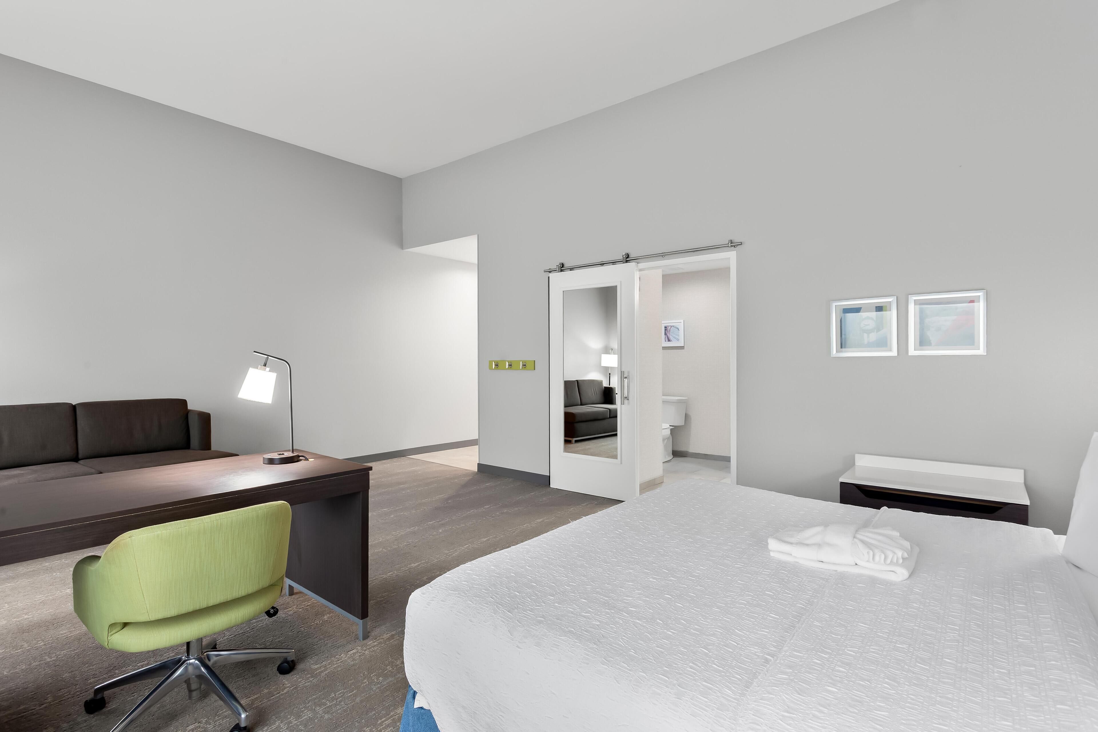 A modern hotel room with a large bed, a desk, a green office chair, a bedside table, and a walk-in closet.