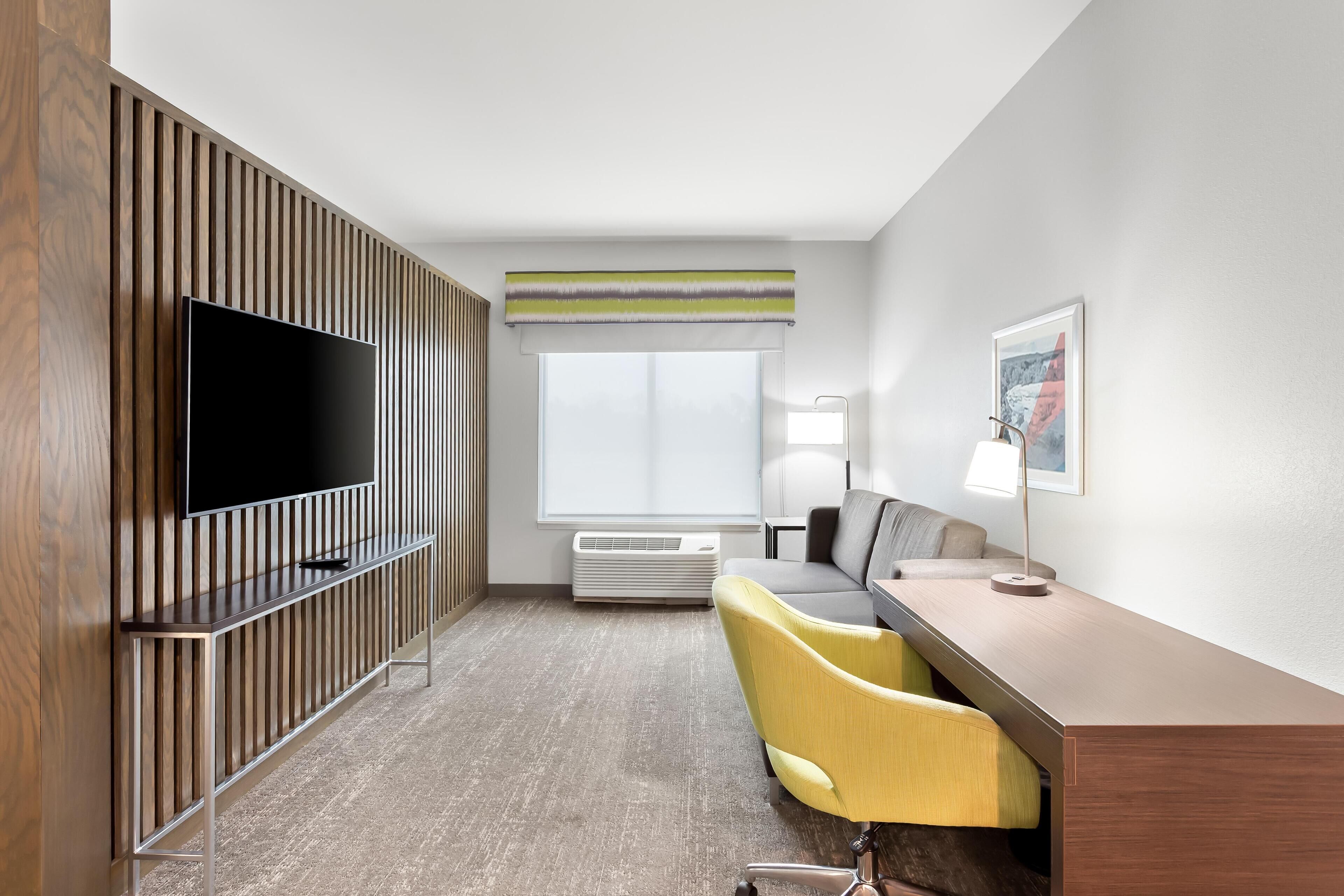A modern hotel room with a flat-screen TV, a wooden panel wall, a large window with a view, a desk with a chair, and a lamp.
