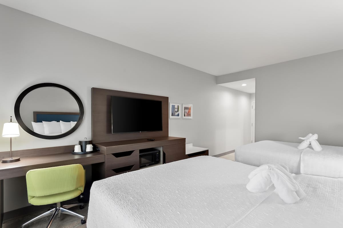 Quarto de hotel com cama, escrivaninha com computador, televisão, espelho e mesinha de cabeceira com abajur.