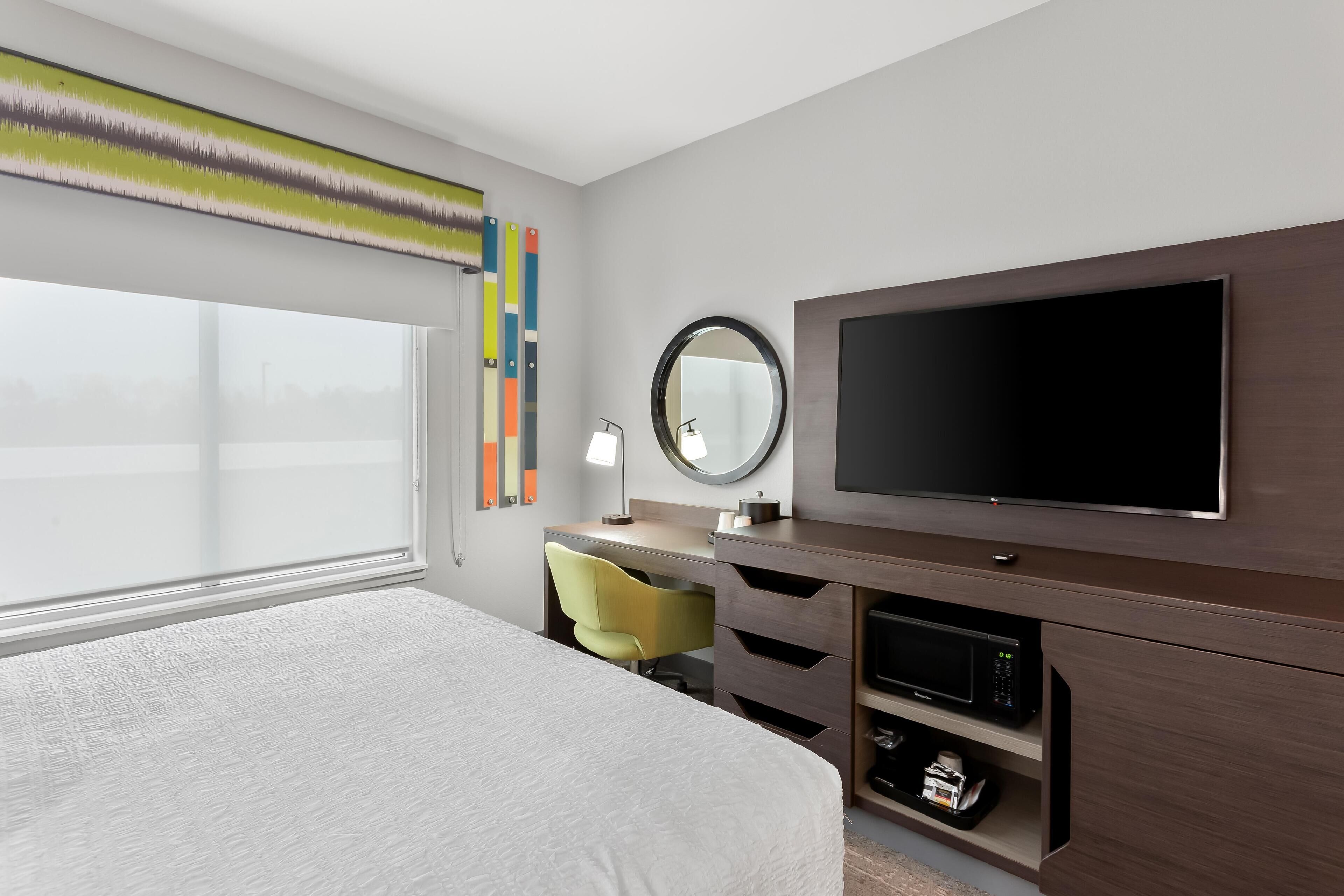 A hotel room with a bed, a desk with a chair, a television, a mirror, and a window.
