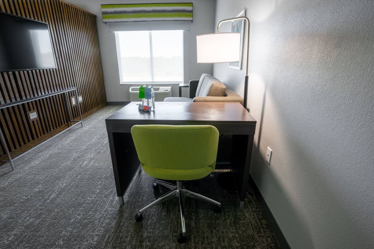 Quarto de hotel com uma escrivaninha, uma cadeira de escritório verde, uma televisão, um abajur e uma janela com persianas.