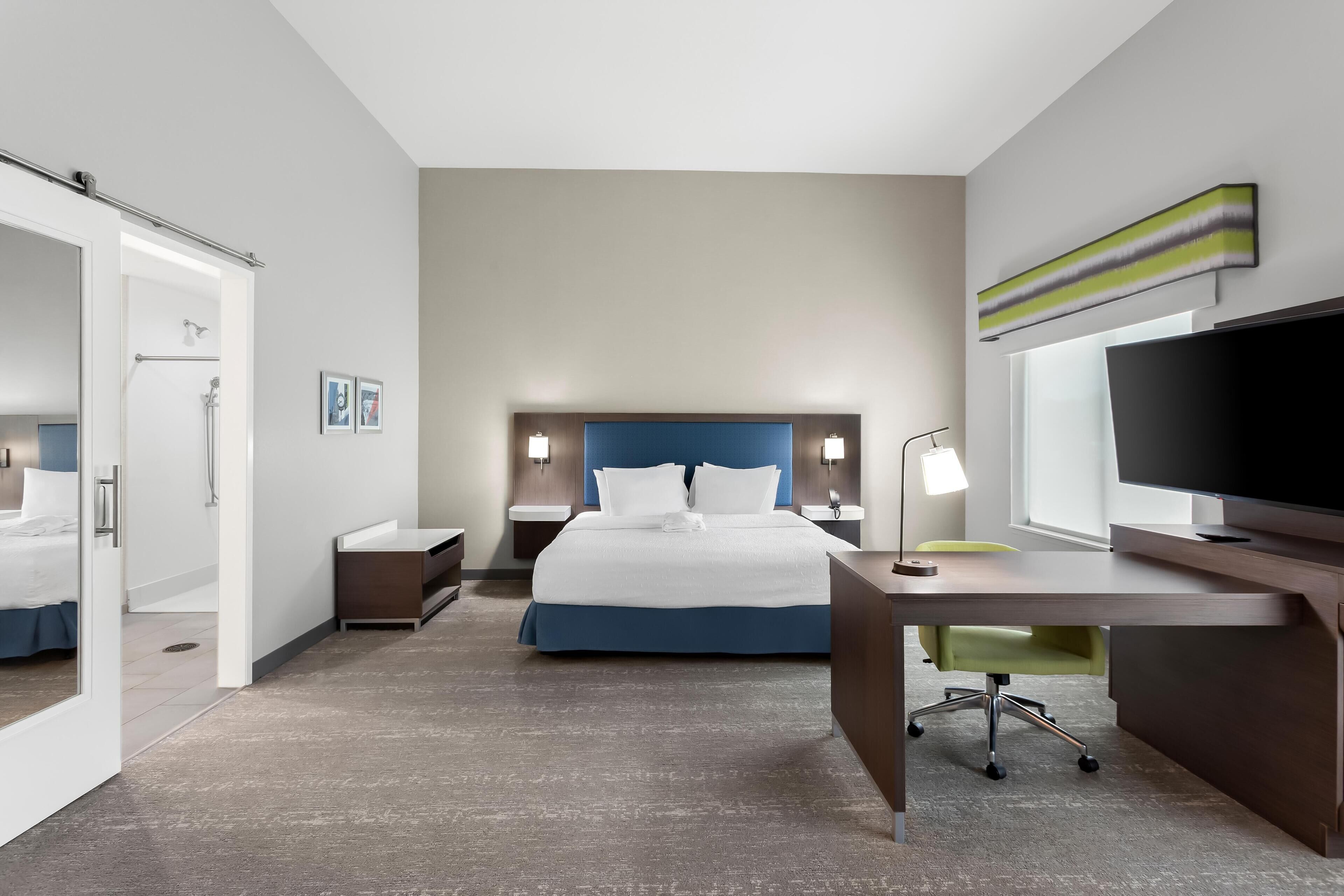 A modern hotel room with a large bed, a desk with a chair, a television, and a bathroom with a shower and toilet.